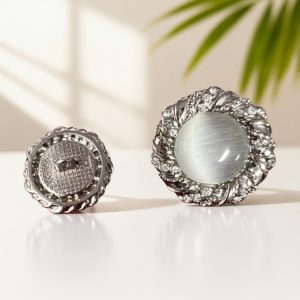 Two silver-tone rhinestone cat eye buttons on a cream background, one turned to show the shank back and one showing the grey cat eye cabochon inside a twisted rhinestone frame
