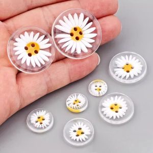 A hand holding two large clear resin daisy buttons with five smaller sizes laid out below, showing how small the daisy buttons are in real terms