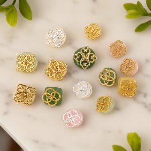 Jade filigree buttons arranged on a marble surface with green leaves, faux jade glass domes in white, yellow, green and pink with openwork gold-tone and silver-tone metalwork, in filigree and clover styles