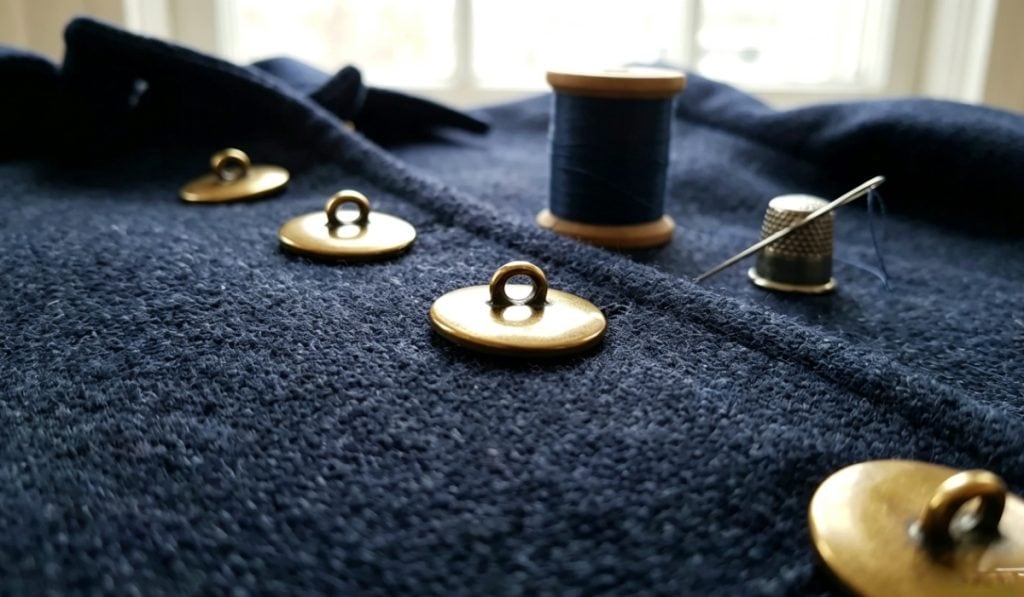 Gold-tone metal shank buttons sewn down the front of a navy wool coat, with a spool of thread, a needle and a thimble resting on the fabric