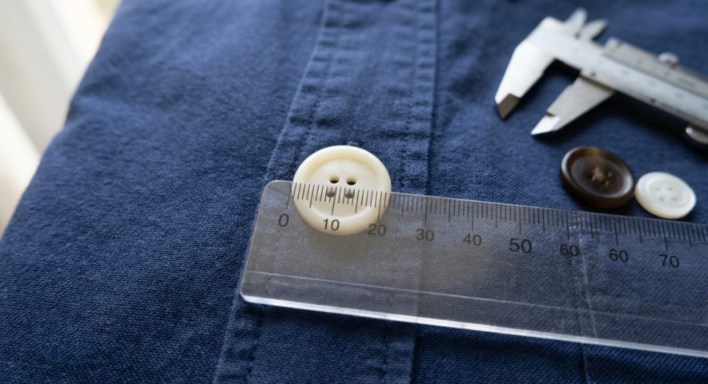 A millimeter ruler laid across the face of a cream four-hole button on a navy shirt to measure its diameter for a replacement