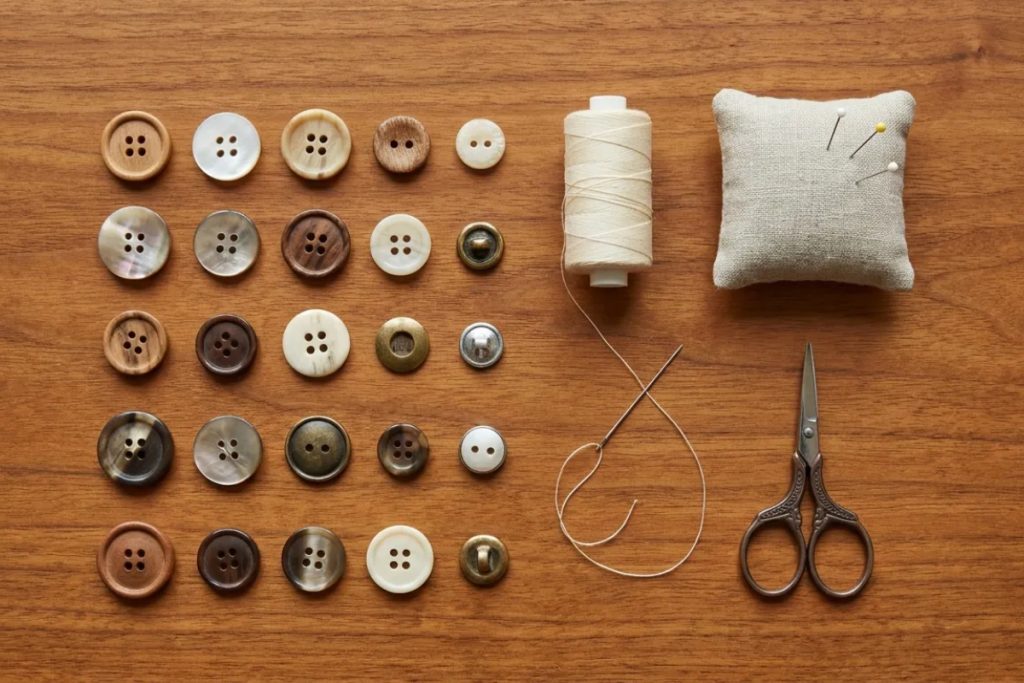A basic hand-sewing kit with assorted buttons, thread, a needle, pincushion and scissors laid out on a wooden surface