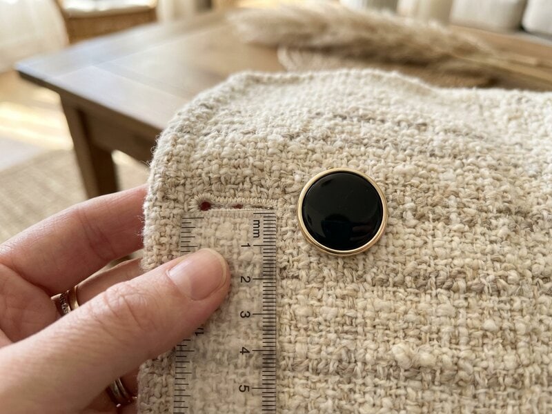 Measuring the buttonhole on a cream tweed jacket with a millimeter ruler next to a black enamel gold-tone shank button