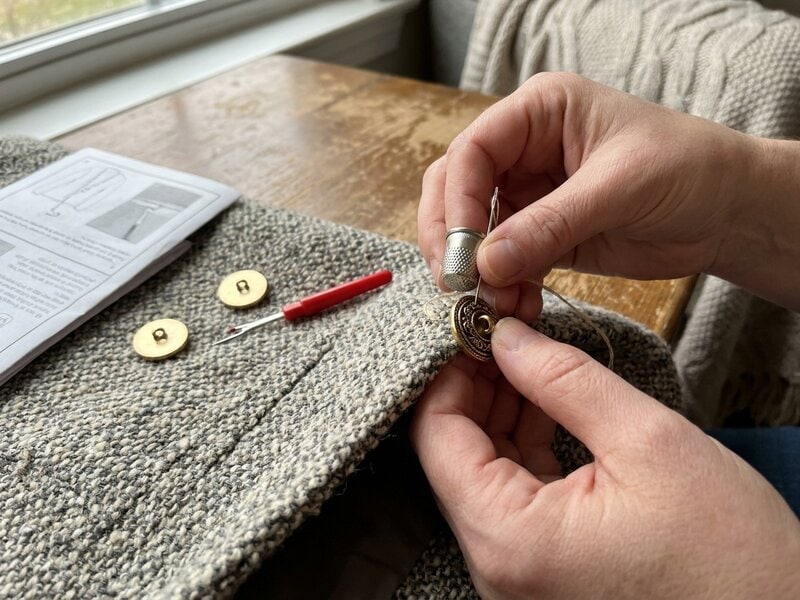 Hands sewing a gold-tone shank button onto the edge of a grey tweed jacket with a needle, thread and seam ripper