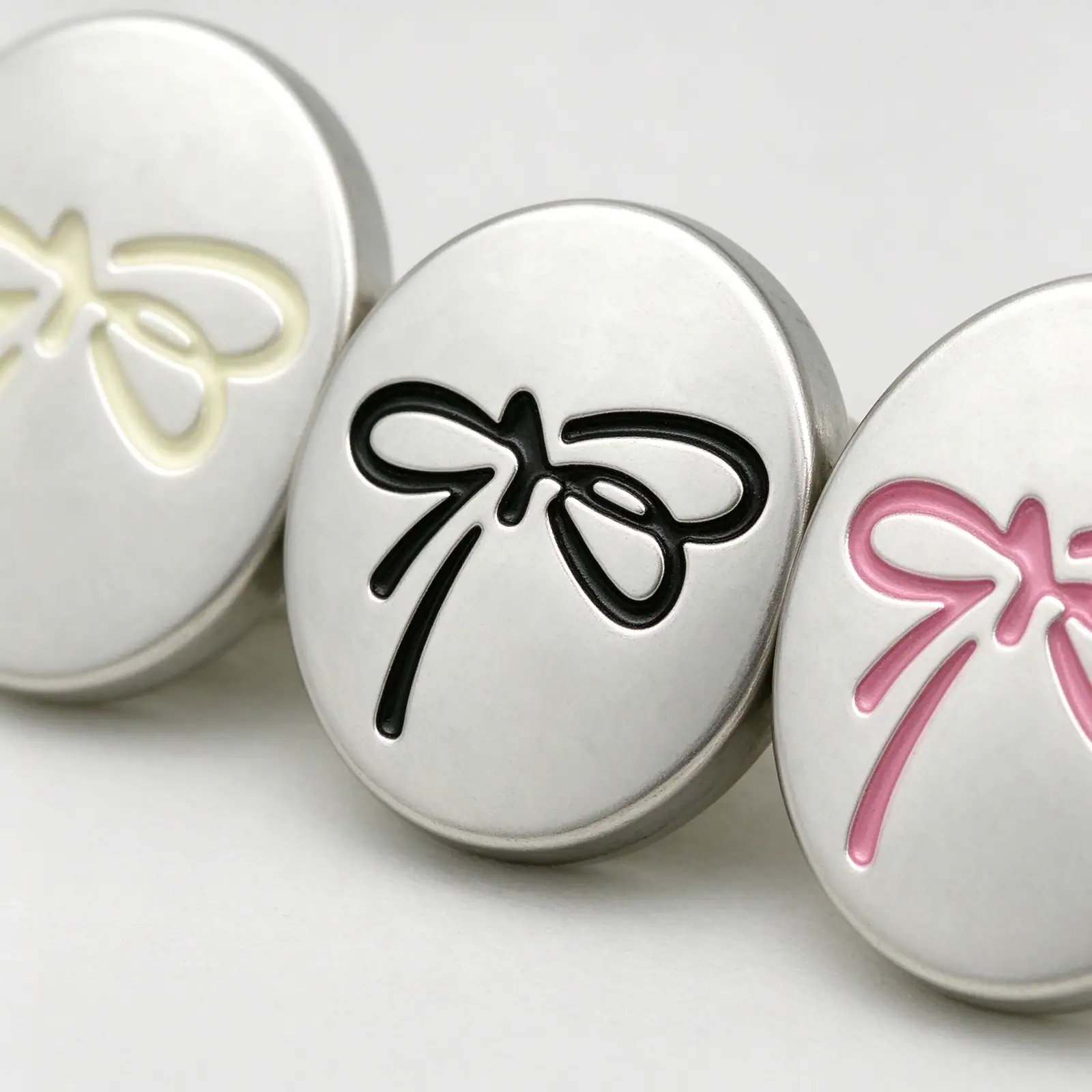 Macro view of three silver bow buttons showing the raised cream, black and pink bow lines on a matte silver face