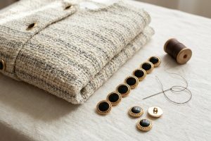 Tweed jacket buttons in gold-tone enamel with braided rims laid out beside a folded cream and grey boucle jacket with thread and needle